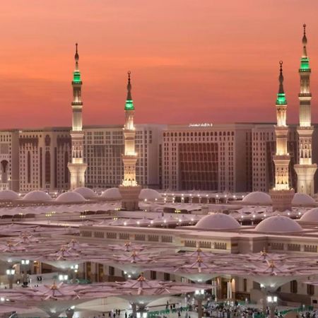 Intercontinental Dar Al Iman Madinah By Ihg Hotel Медина Екстериор снимка The photo depicts a scenic view of a mosque complex, likely during sunset, with several minarets illuminated and showcasing green lights. The architecture features domed roofs, and the setting includes a backdrop of buildings, all bathed in a warm, o