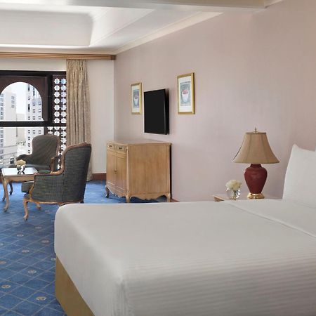 Intercontinental Dar Al Iman Madinah By Ihg Hotel Медина Екстериор снимка The photo shows a well-furnished hotel room. It features a large bed with a white duvet, two bedside lamps, and decorative paintings on the walls. There is a comfortable seating area with two chairs and a small table. A wooden dresser is positioned a
