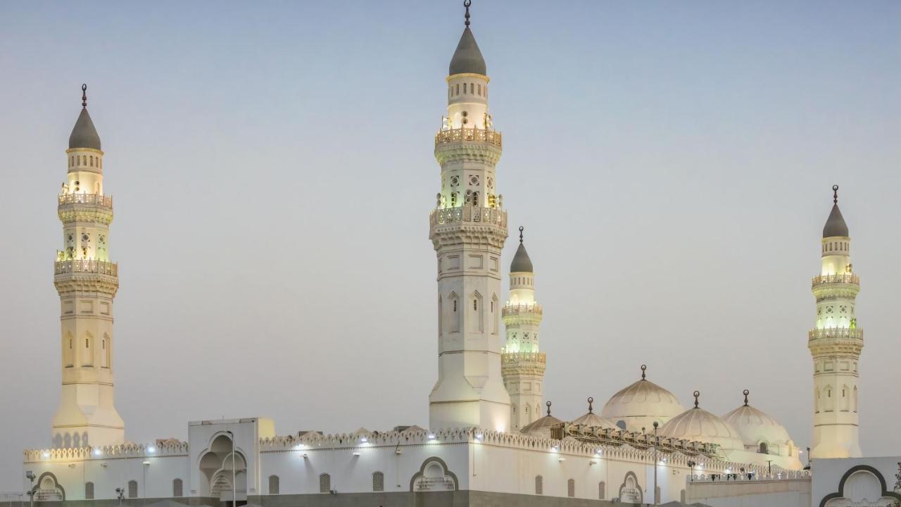 Intercontinental Dar Al Iman Madinah By Ihg Hotel Медина Екстериор снимка The image shows a mosque with distinct architectural features, including tall minarets and domes. The minarets are highlighted, likely lit up in the evening, showcasing intricate design elements typical of Islamic architecture. The overall scene conv