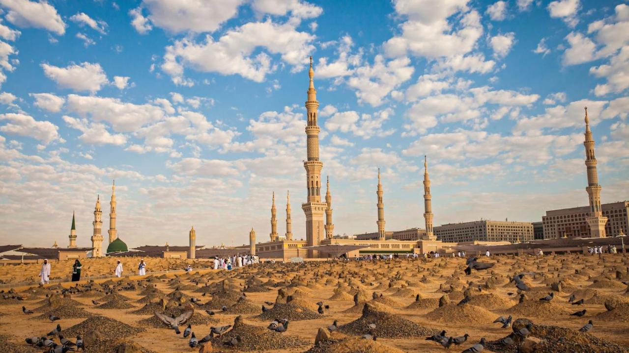 Intercontinental Dar Al Iman Madinah By Ihg Hotel Медина Екстериор снимка The photo shows a landscape that features a large mosque with a tall minaret surrounded by smaller minarets. In the foreground, there are numerous grave markers arranged in a desert-like setting. The sky is bright with scattered clouds, and there are