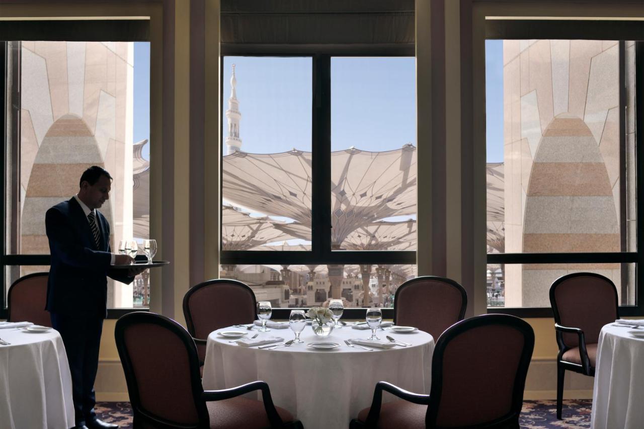 Intercontinental Dar Al Iman Madinah By Ihg Hotel Медина Екстериор снимка The photo shows a dining area with elegant decor. A waiter is standing to the side, holding a tray. The tables are neatly set with white tablecloths and fine dining arrangements. Large windows provide a view of an outdoor area featuring architectural