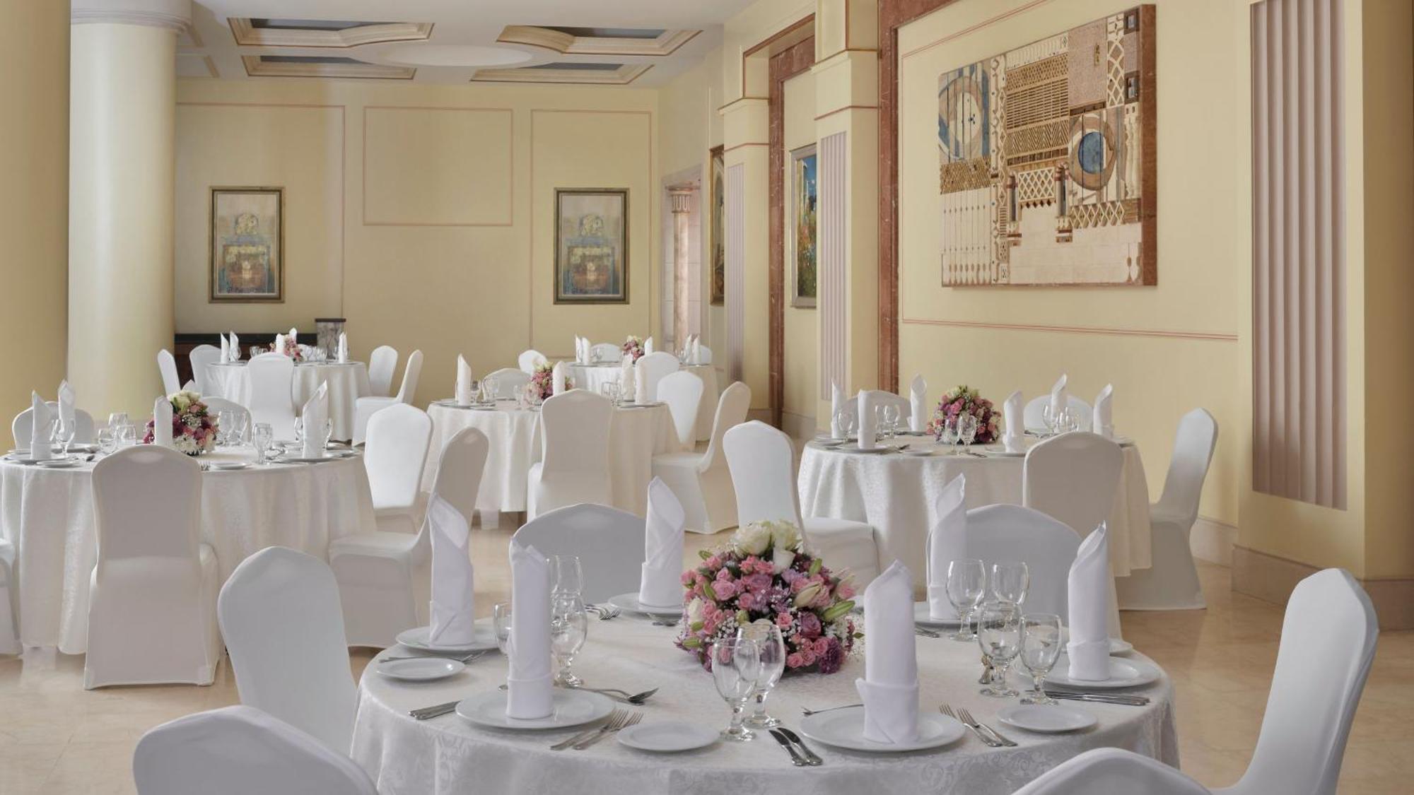Intercontinental Dar Al Iman Madinah By Ihg Hotel Медина Екстериор снимка The photo shows an elegantly arranged dining area, likely set up for a formal event or reception. There are several round tables covered with white tablecloths, each adorned with floral centerpieces featuring pink flowers. The tables are set with pla