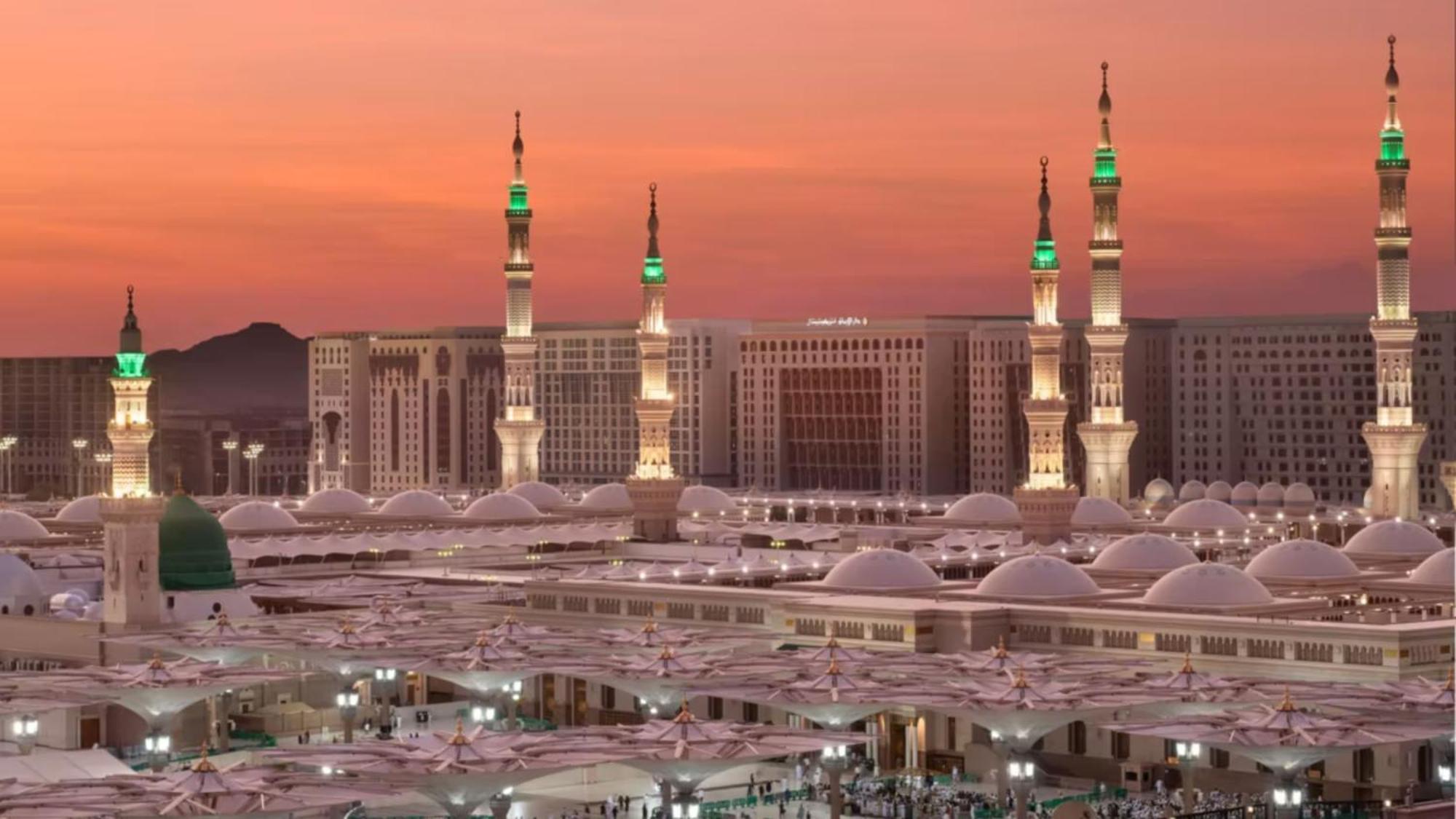 Intercontinental Dar Al Iman Madinah By Ihg Hotel Медина Екстериор снимка The photo depicts a scenic view of a mosque complex, likely during sunset, with several minarets illuminated and showcasing green lights. The architecture features domed roofs, and the setting includes a backdrop of buildings, all bathed in a warm, o