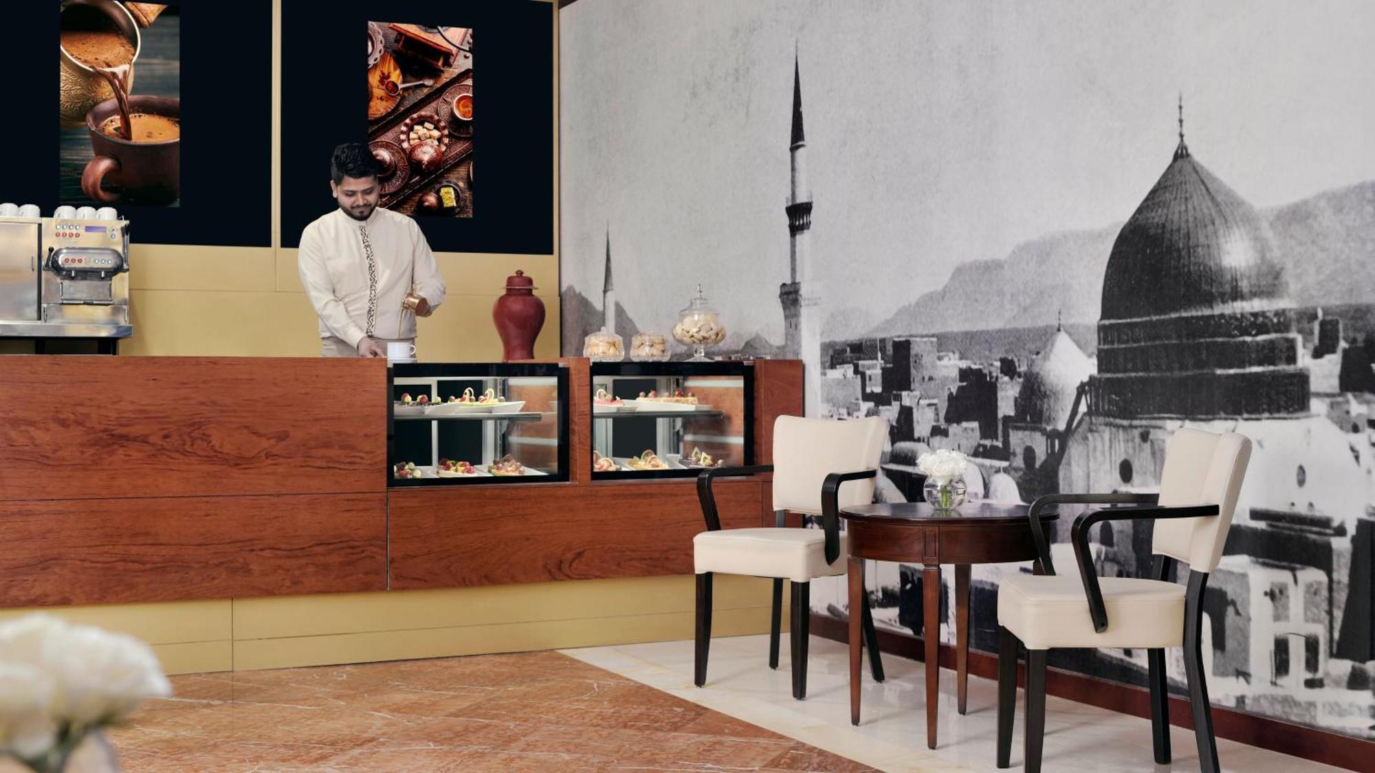 Intercontinental Dar Al Iman Madinah By Ihg Hotel Медина Екстериор снимка The photo shows an elegant cafe or dessert shop interior. A man in traditional attire is standing behind a wooden counter, where various pastries and sweets are displayed. The walls feature a large black-and-white mural of a cityscape, possibly depic