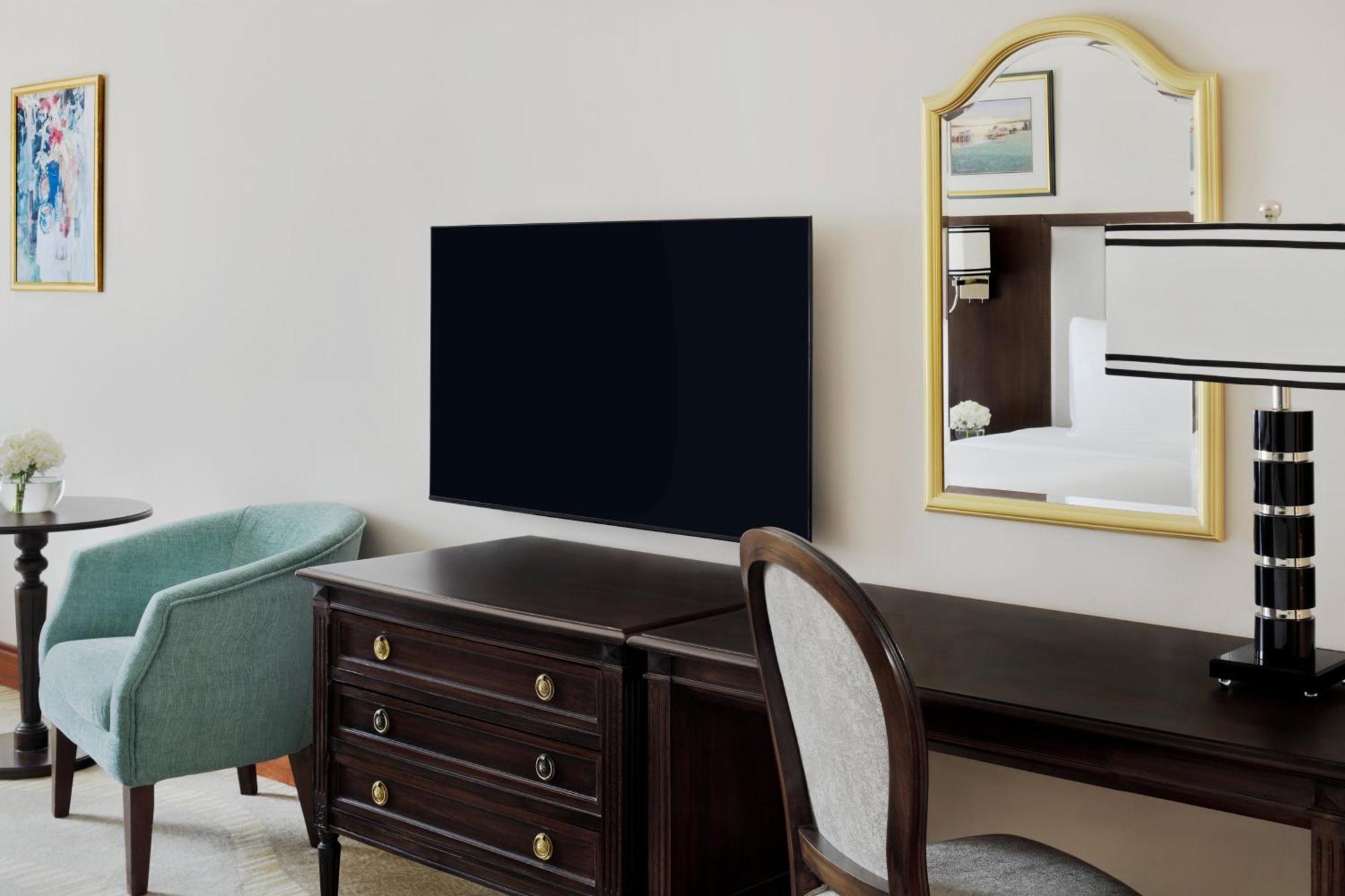 Intercontinental Dar Al Iman Madinah By Ihg Hotel Медина Екстериор снимка The photo shows a hotel room interior featuring a mounted flat-screen television on the wall. Below the TV, there is a dark wooden dresser with drawers. To the right, there's a wooden desk and a chair with a light-colored seat. A small table with a l
