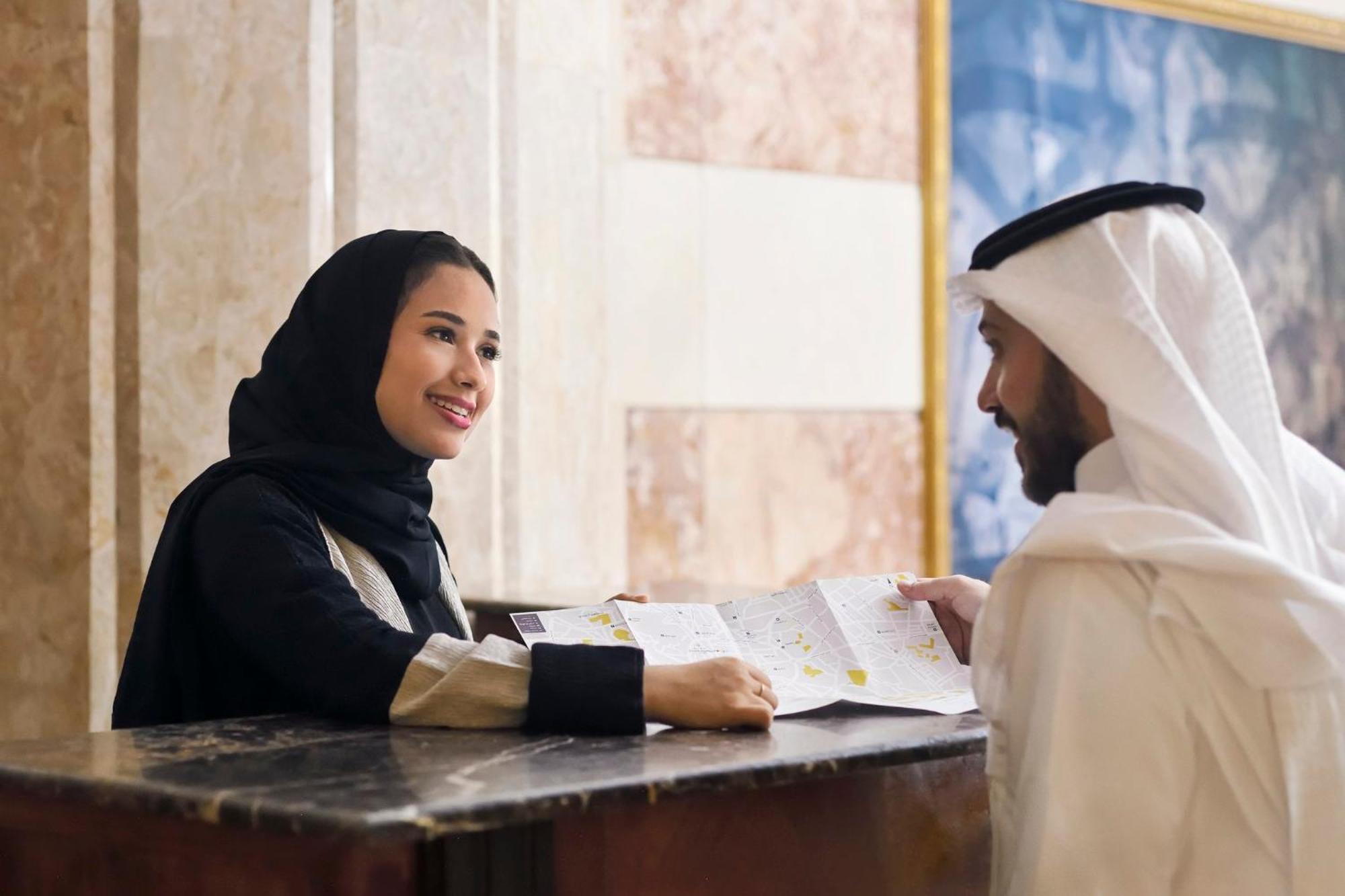 Intercontinental Dar Al Iman Madinah By Ihg Hotel Медина Екстериор снимка The photo depicts a woman in a black hijab and a light-colored outfit smiling as she interacts with a man wearing traditional Emirati attire, including a white kandura and ghutrah (headscarf). They are at a reception desk in what appears to be a hote