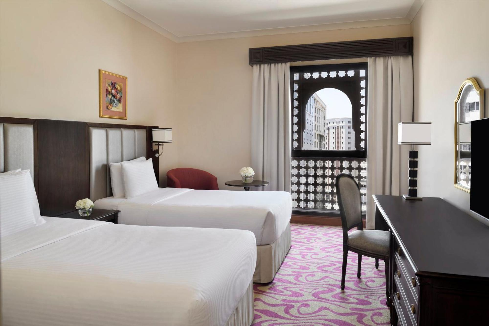 Intercontinental Dar Al Iman Madinah By Ihg Hotel Медина Екстериор снимка The photo shows a hotel room featuring two single beds with white bedding. The room has a light, neutral color scheme and is decorated with minimal furnishings. There's a small seating area with a red chair and a round table, as well as a desk with a