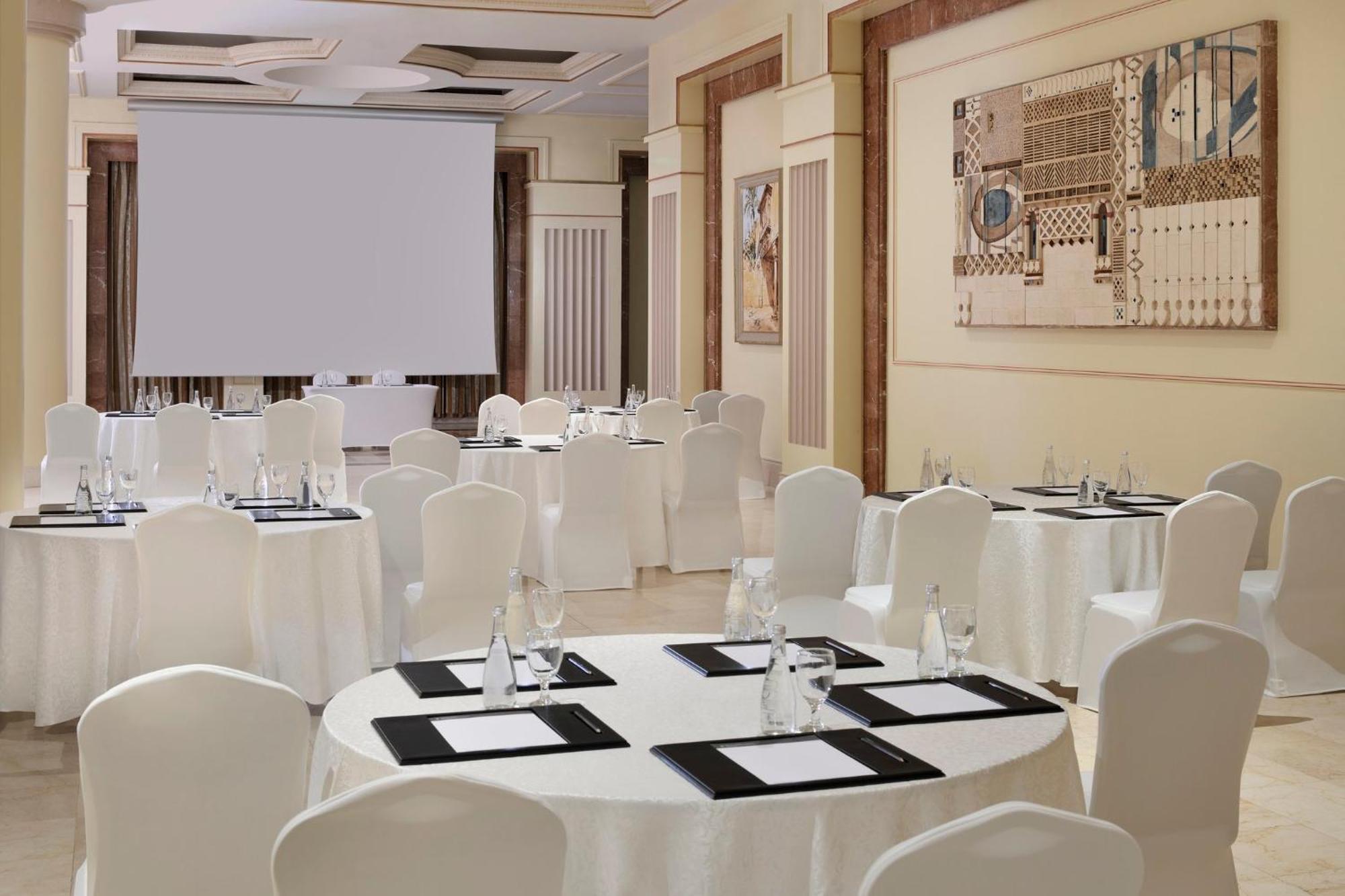 Intercontinental Dar Al Iman Madinah By Ihg Hotel Медина Екстериор снимка The photo shows a well-decorated event space or meeting room. It features several round tables covered with white tablecloths and surrounded by white chairs. Each table has black placemats and glass water bottles, suggesting a formal setup for a gath