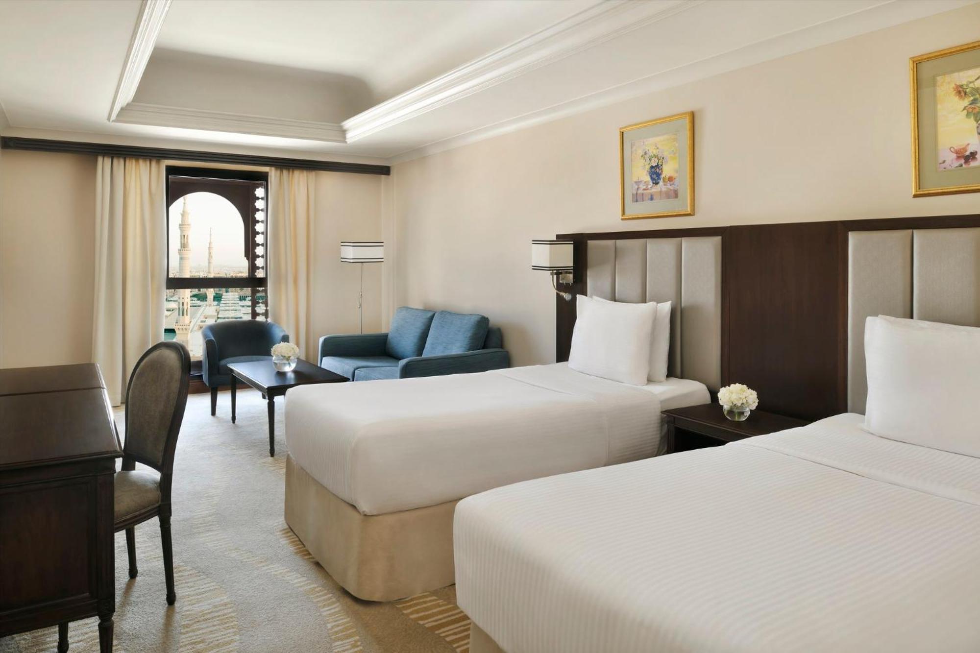 Intercontinental Dar Al Iman Madinah By Ihg Hotel Медина Екстериор снимка The photo shows a well-appointed hotel room featuring two twin beds with white bedding. There is a small desk with a chair, a comfortable blue sofa, and a coffee table in the sitting area. The room has a warm, neutral color scheme, accented by two pa