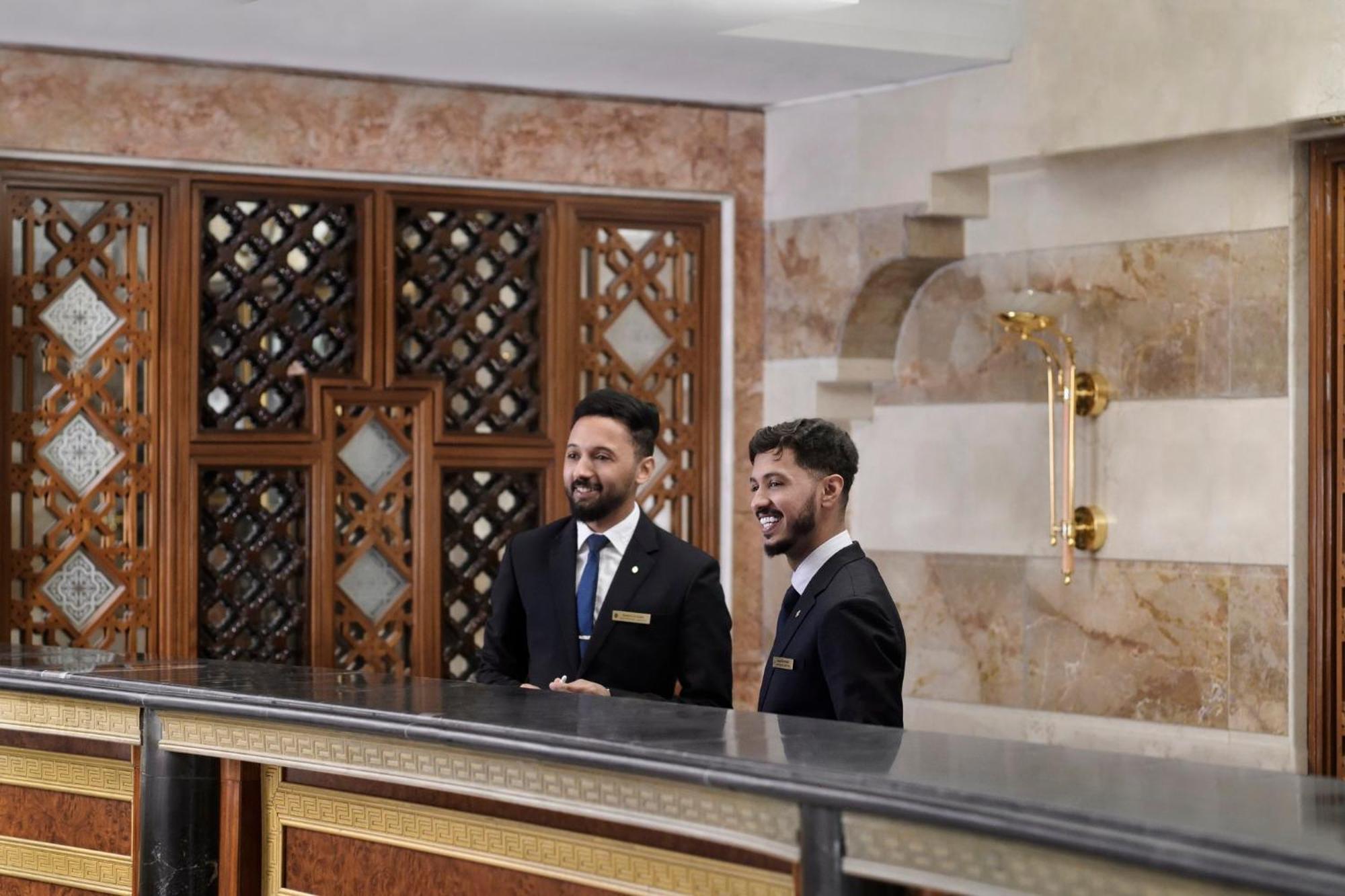 Intercontinental Dar Al Iman Madinah By Ihg Hotel Медина Екстериор снимка The photo shows two men standing behind a reception desk in an elegant setting, likely a hotel or similar establishment. They are both dressed in formal attire, wearing suits and ties, and appear to be smiling or engaging in conversation. The backgro
