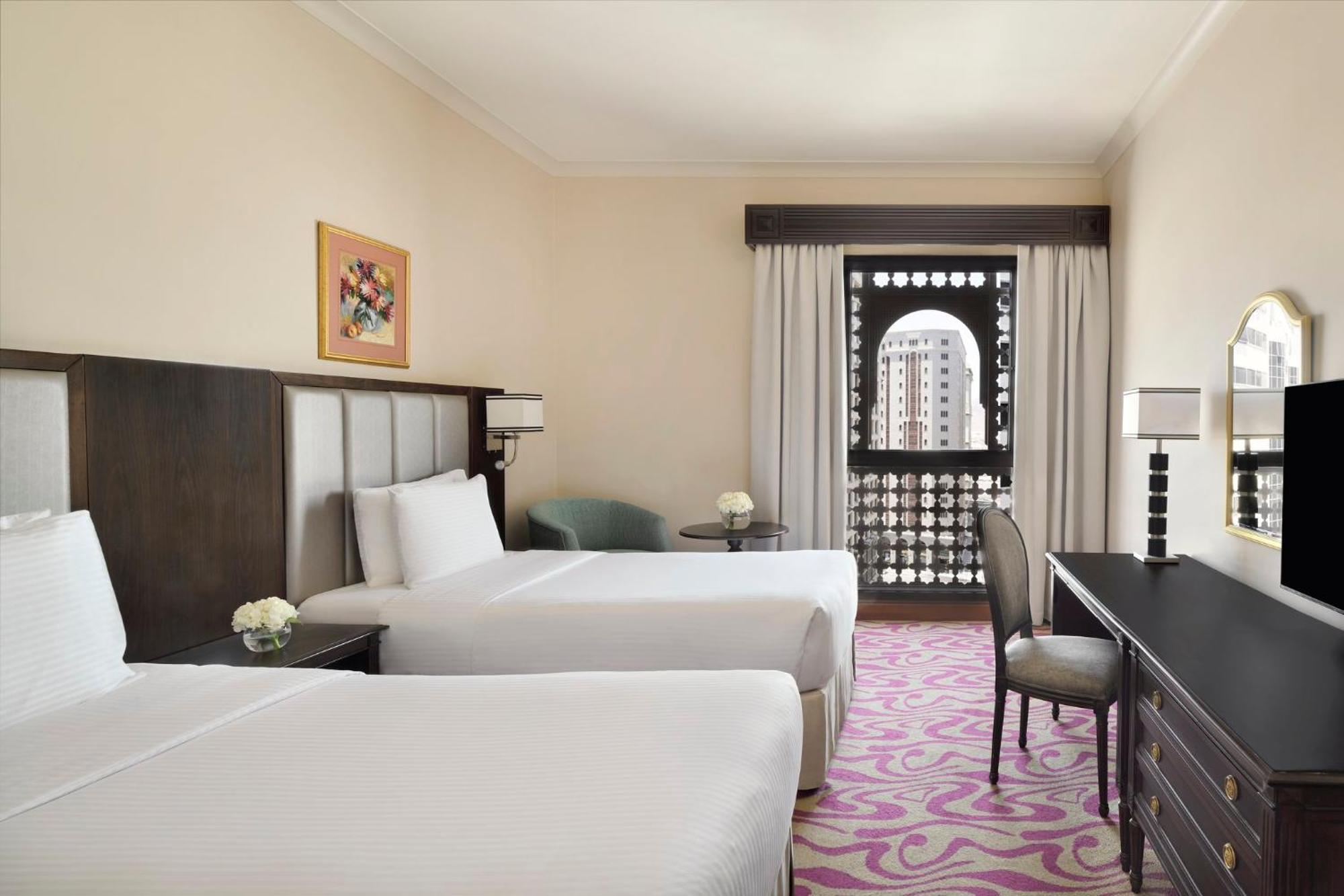 Intercontinental Dar Al Iman Madinah By Ihg Hotel Медина Екстериор снимка The photo shows a hotel room featuring two twin beds with white bedding. There is a decorative piece of art on the wall above one of the beds. A small round table with a flower arrangement is placed next to the beds. A desk and chair are situated aga