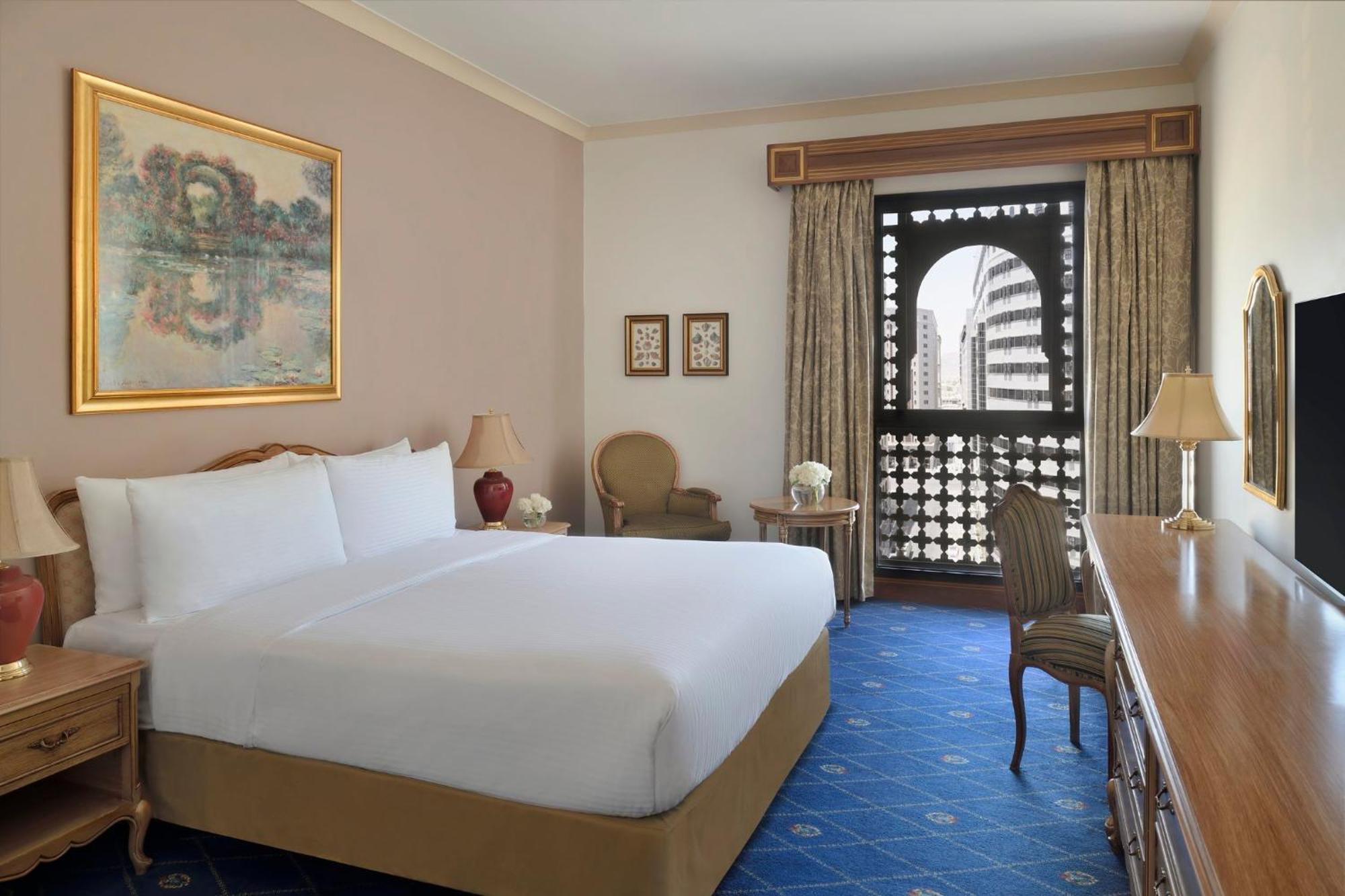 Intercontinental Dar Al Iman Madinah By Ihg Hotel Медина Екстериор снимка The photo shows a hotel room that features a large, neatly made bed with white linens. There is a framed painting on the wall above the bed. To the side, a small seating area includes a chair and a side table. Opposite the bed, there is a wooden desk