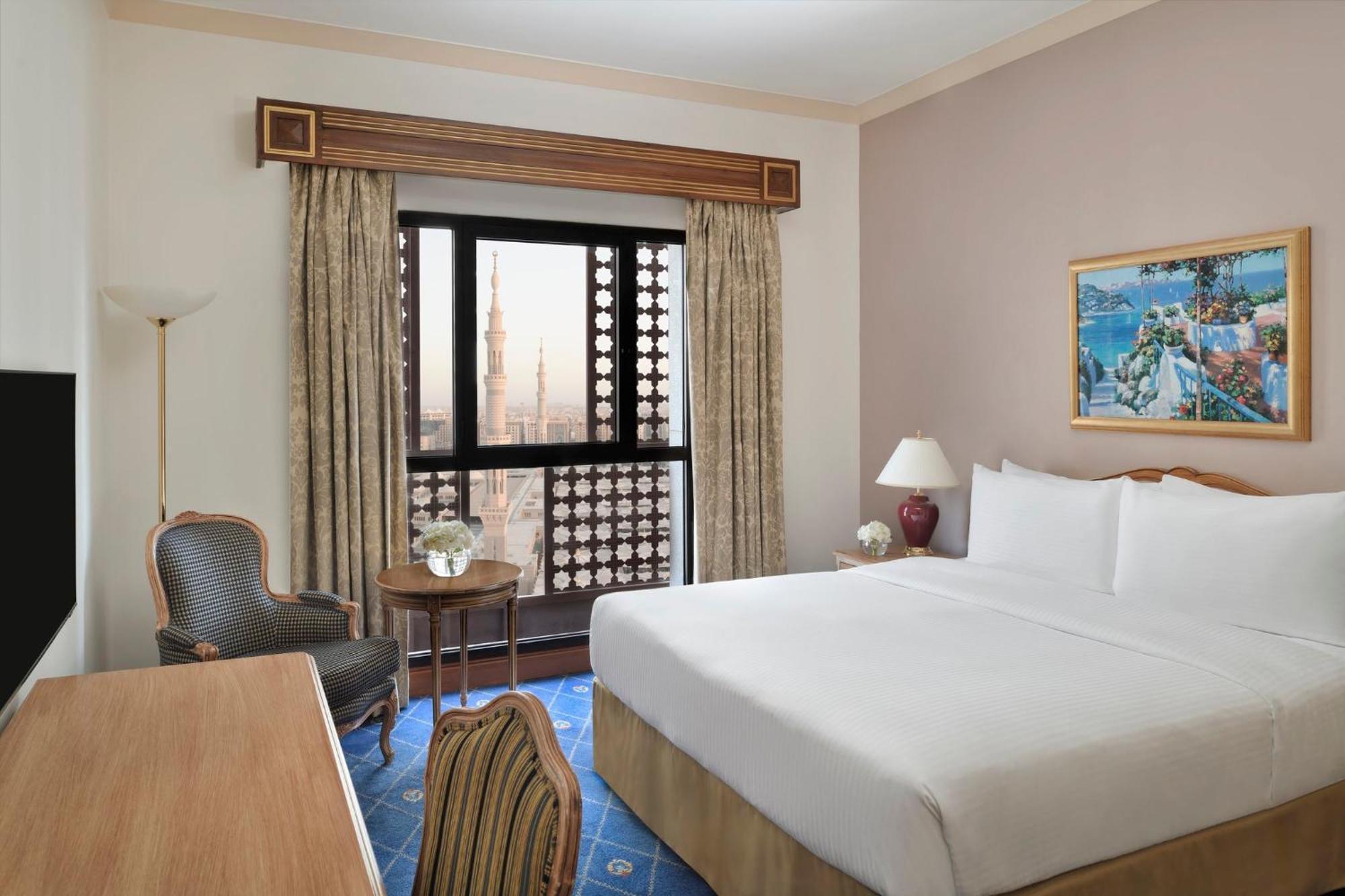 Intercontinental Dar Al Iman Madinah By Ihg Hotel Медина Екстериор снимка The photo shows a hotel room with a modern design. There is a large bed with white linens placed against the wall. Next to the bed, there is a small bedside table with a lamp. A comfortable armchair is situated nearby, accompanied by a wooden desk. T