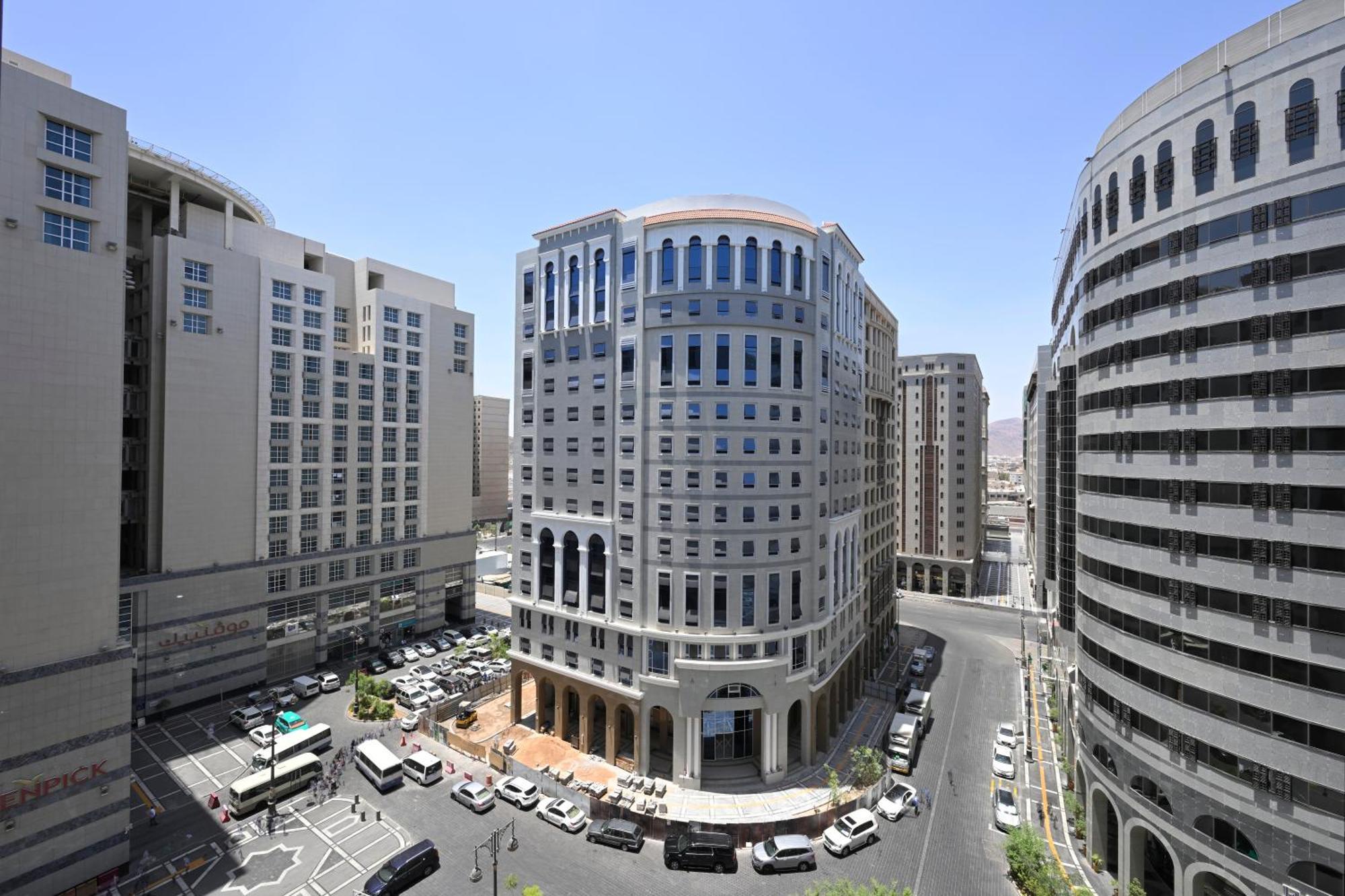 Intercontinental Dar Al Iman Madinah By Ihg Hotel Медина Екстериор снимка The photo shows a modern urban scene featuring several buildings, including a prominent rounded building with a glass façade. This building appears to be situated at an intersection, surrounded by other high-rise structures that have distinct archite