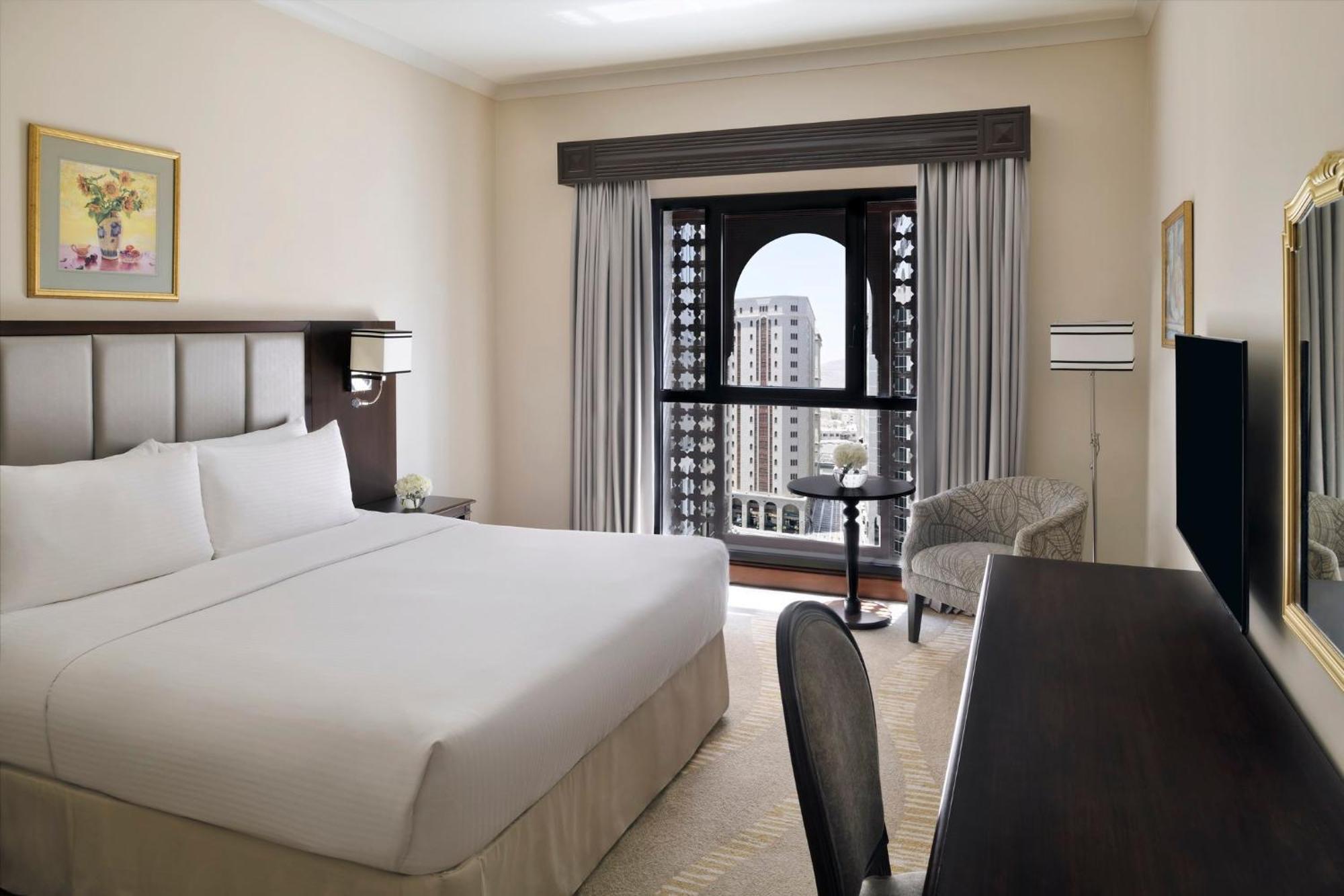 Intercontinental Dar Al Iman Madinah By Ihg Hotel Медина Екстериор снимка The photo shows a hotel room with a modern and minimalistic design. It features a large bed with white linens, a side table, and a comfortable chair near a window. The window has a view of a cityscape, and elegant curtains frame the window. There is 