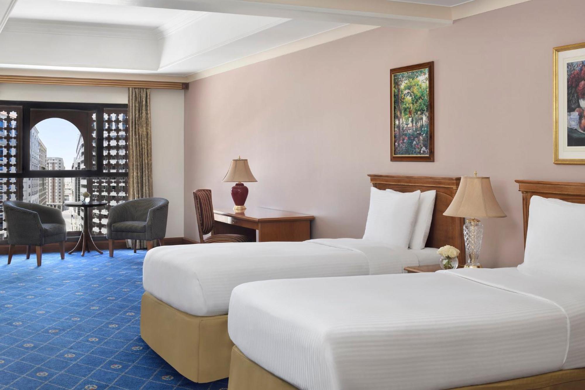 Intercontinental Dar Al Iman Madinah By Ihg Hotel Медина Екстериор снимка The photo shows a nicely designed hotel room featuring two twin beds with white linens and beige bases. In the background, there's a wooden desk and a lamp, along with a pair of comfortable armchairs positioned near a large window. The window showcas