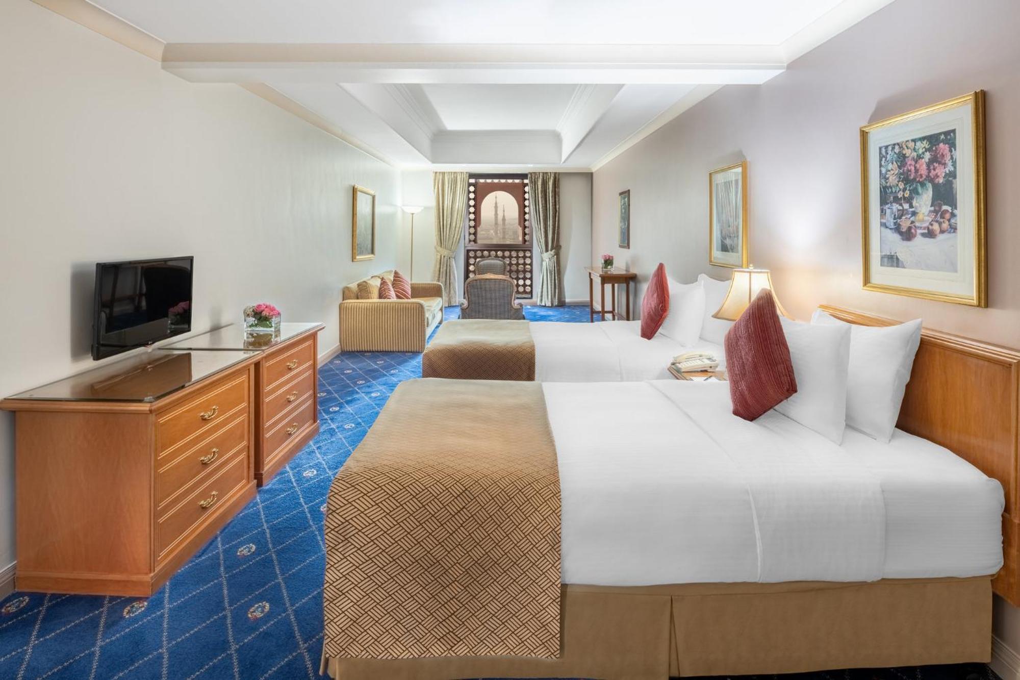 Intercontinental Dar Al Iman Madinah By Ihg Hotel Медина Екстериор снимка The photo shows a hotel room featuring two beds with white linens and decorative pillows. There is a wooden dresser with a television on it. The flooring has a blue carpet with a pattern, and the walls are painted in a light color. The room is well-l