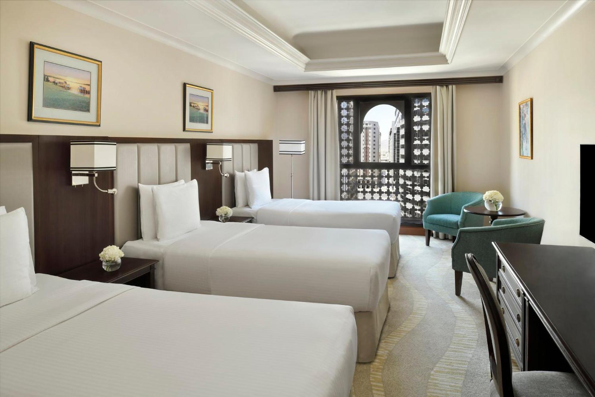 Intercontinental Dar Al Iman Madinah By Ihg Hotel Медина Екстериор снимка The photo shows a hotel room with three single beds, dressed in white linens. There are two paintings on the walls, depicting serene landscapes. A large window allows natural light to enter, and it is adorned with elegant curtains. A smaller seating 