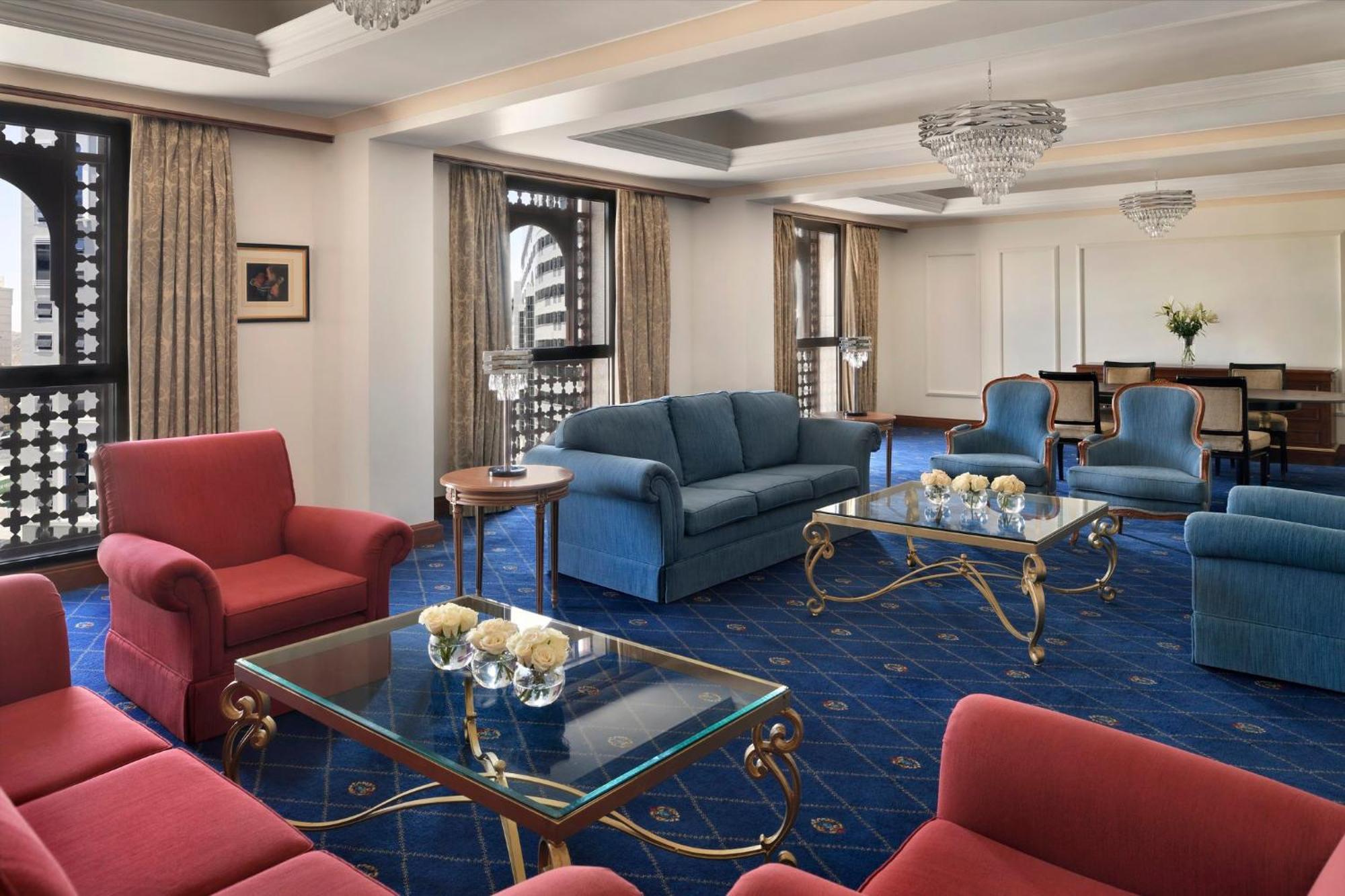 Intercontinental Dar Al Iman Madinah By Ihg Hotel Медина Екстериор снимка The photo shows a luxurious living space or lounge area. It features a combination of furniture, including a blue sofa, several red armchairs, and glass-top coffee tables with gold accents. The flooring is carpeted in a deep blue, and the walls are a