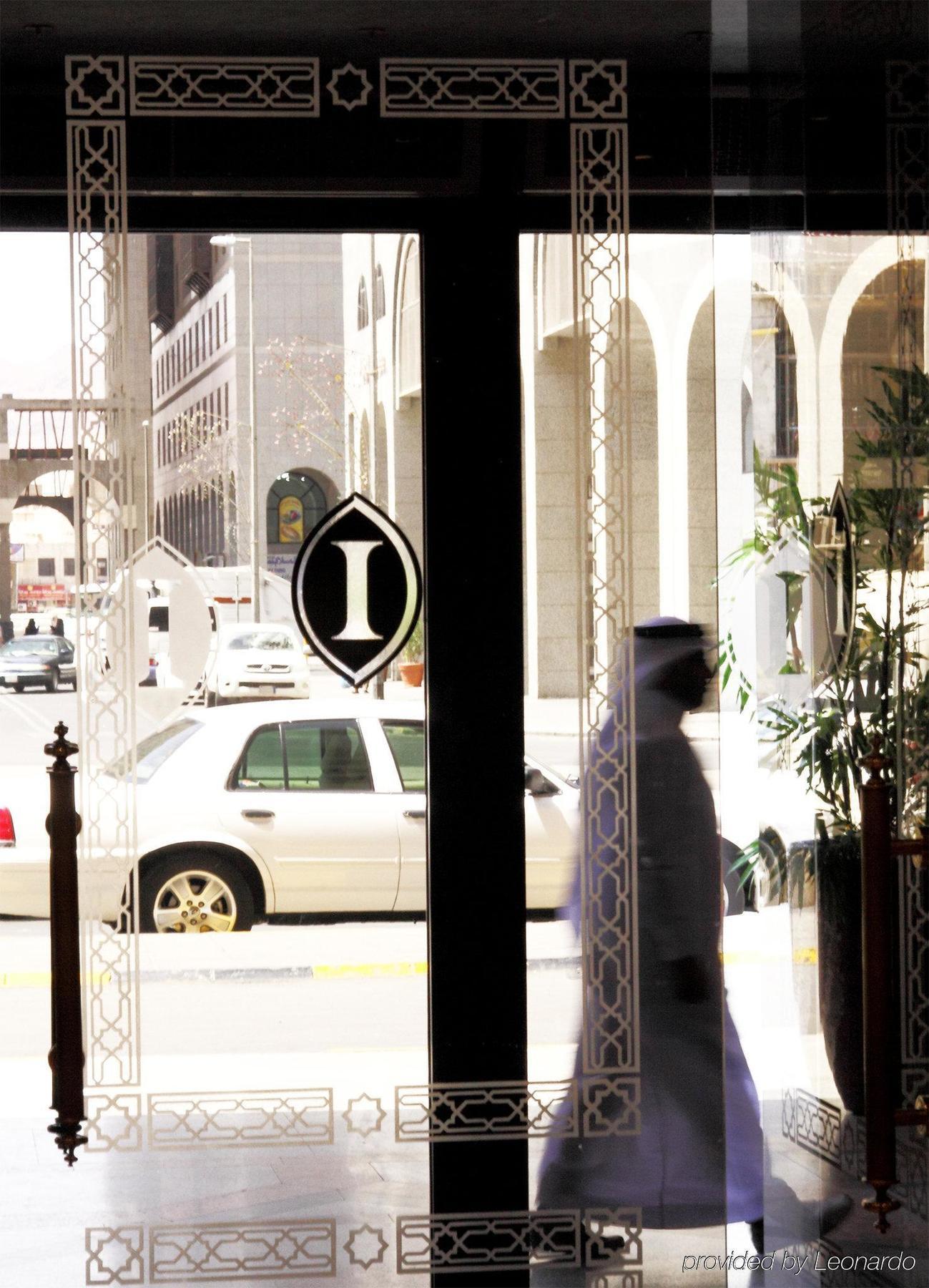 Intercontinental Dar Al Iman Madinah By Ihg Hotel Медина Интериор снимка The photo shows a view through a glass door or window, featuring an elegant design with a prominent "I" symbol displayed. Through the glass, there are some parked vehicles visible, including a white car. In the foreground, a shadowy figure of a perso