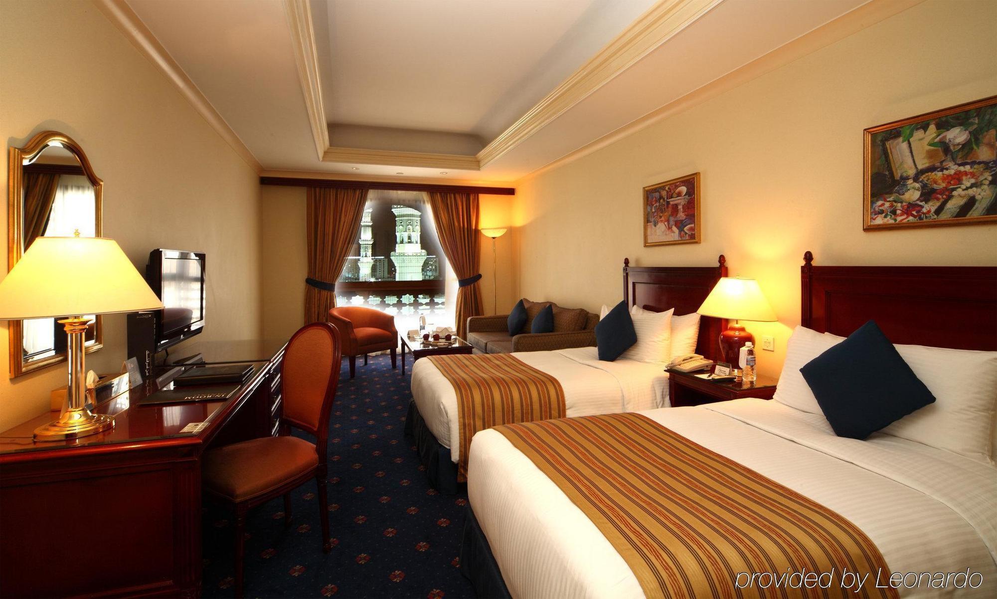 Intercontinental Dar Al Iman Madinah By Ihg Hotel Медина Стая снимка The photo shows a hotel room featuring two beds with white linens and brown striped throws. There is a wooden desk and chair, alongside a television. The walls are painted in a warm tone, and there are decorative paintings. Large windows with curtain