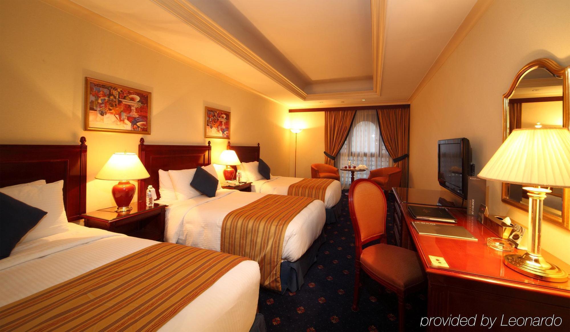 Intercontinental Dar Al Iman Madinah By Ihg Hotel Медина Стая снимка The photo shows a hotel room with a warm and inviting atmosphere. There are two beds with white and gold bedding, complemented by decorative pillows. Each bed has a bedside lamp. In the background, there's a small seating area with a couple of chairs