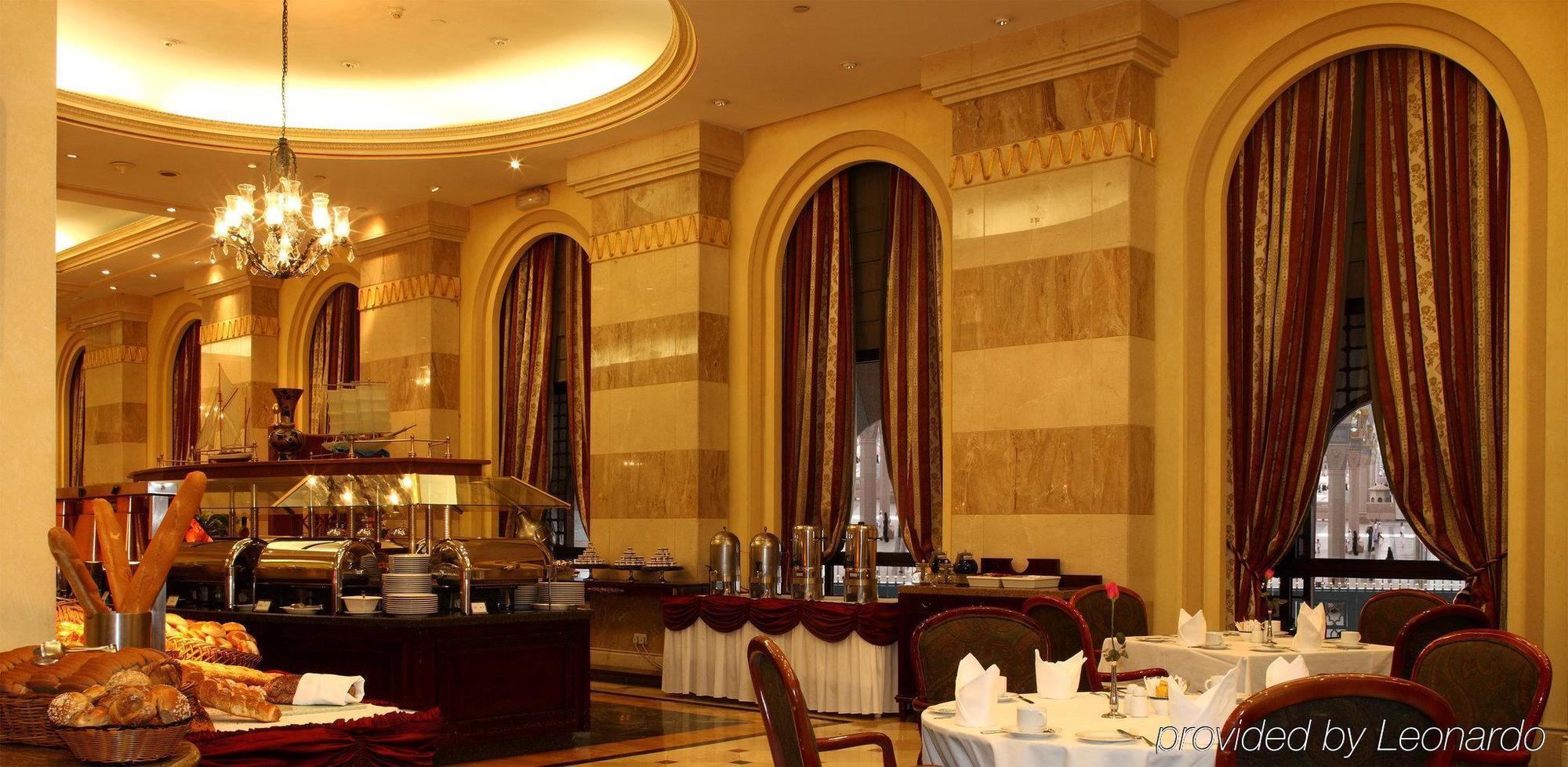 Intercontinental Dar Al Iman Madinah By Ihg Hotel Медина Ресторант снимка The photo shows an elegant dining restaurant interior. It features stylish arches and walls with a blend of marble and decorative elements. There are large windows draped with rich curtains, contributing to the luxurious atmosphere. The dining area i